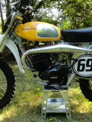 Seitenansicht einer Husqvarna 125CR Lightweight 1974 mit gelbem Tank, im Gelände auf einem Ständer platziert.