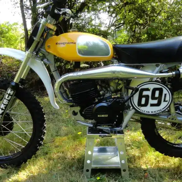 Seitenansicht einer Husqvarna 125CR Lightweight 1974 mit gelbem Tank, im Gelände auf einem Ständer platziert.