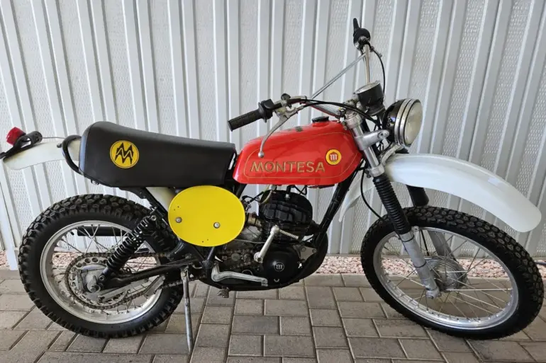 Montesa Enduro 250 von 1975, klassisches Enduro-Motorrad mit rotem Tank und gelben Startnummerntafeln seitlich, vor einer Metallwand abgestellt