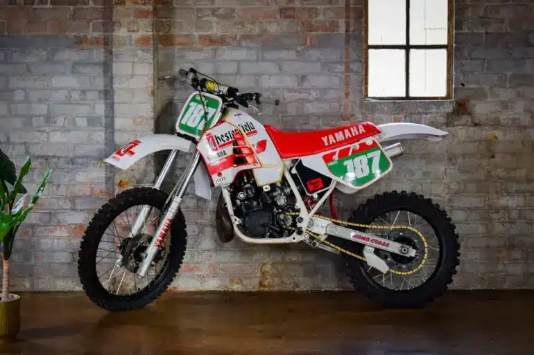 Seitliche Ansicht einer weißen Yamaha YZ250 (Baujahr 1987) mit Chesterfield-Grafiken, abgestellt vor einer Backsteinwand.