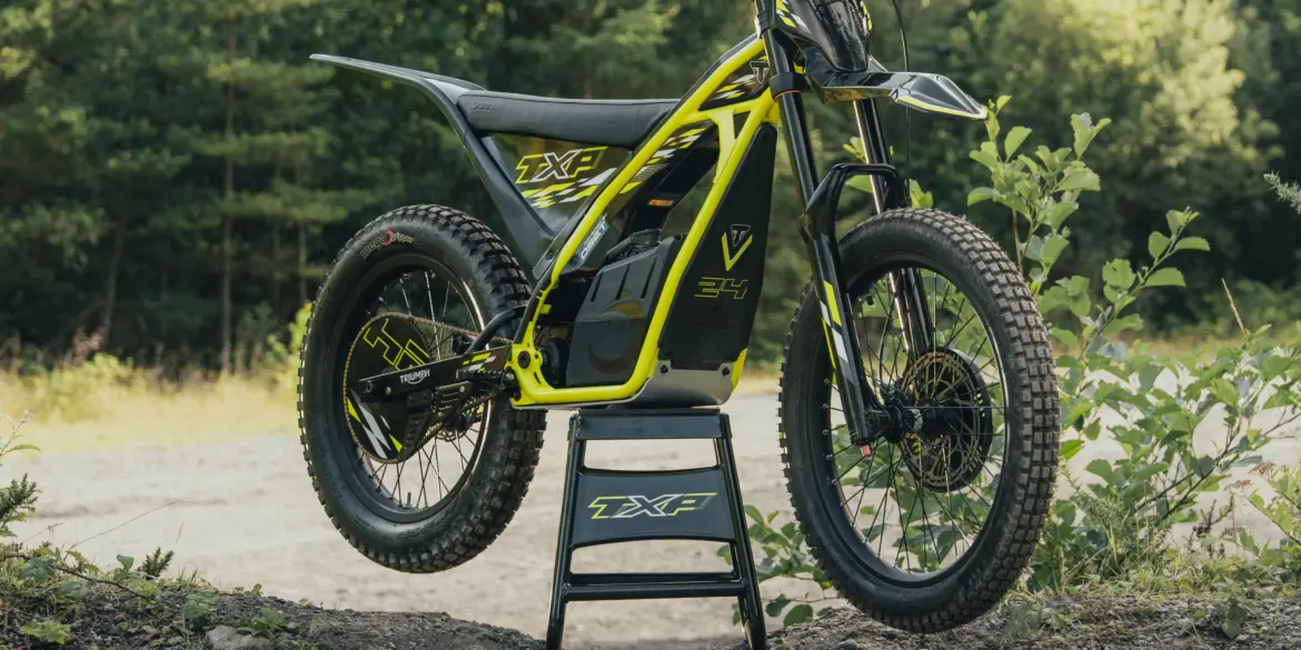 Triumph TXP-24 Elektromotorrad im Offroad-Einsatz auf einem Ständer vor Waldkulisse