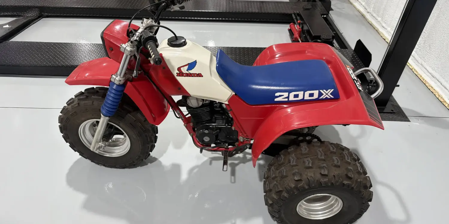 Seitenansicht einer Honda ATC 200X von 1985 mit blauem Sitz, weißem Tank und roten Kunststoffverkleidungen, aufgenommen in einer Werkhalle.