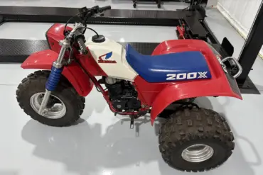 Seitenansicht einer Honda ATC 200X von 1985 mit blauem Sitz, weißem Tank und roten Kunststoffverkleidungen, aufgenommen in einer Werkhalle.