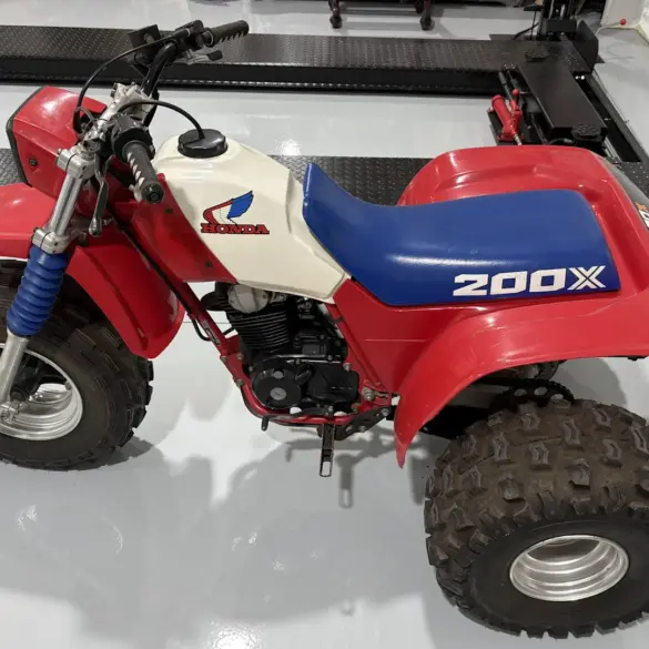 Seitenansicht einer Honda ATC 200X von 1985 mit blauem Sitz, weißem Tank und roten Kunststoffverkleidungen, aufgenommen in einer Werkhalle.