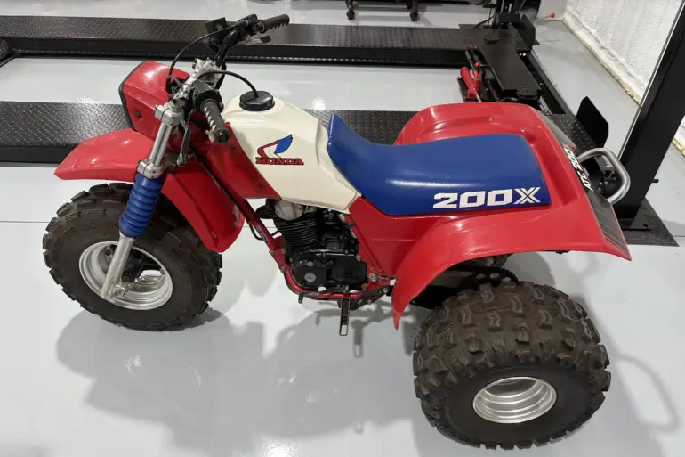 Seitenansicht einer Honda ATC 200X von 1985 mit blauem Sitz, weißem Tank und roten Kunststoffverkleidungen, aufgenommen in einer Werkhalle.
