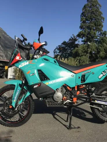 KTM 9X0 Rally Umbau mit hohem Kotflügel, Navigation Tower aus Carbon und schweizerischer Farbgebung, geparkt vor einer Berglandschaft.
