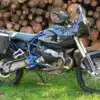 AfricanQueens HP2 Rallye Enduro-Motorrad mit Touratech-Koffern und Carbon-Verkleidung vor Holzstapel.
