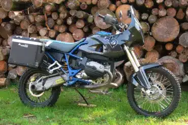 AfricanQueens HP2 Rallye Enduro-Motorrad mit Touratech-Koffern und Carbon-Verkleidung vor Holzstapel.