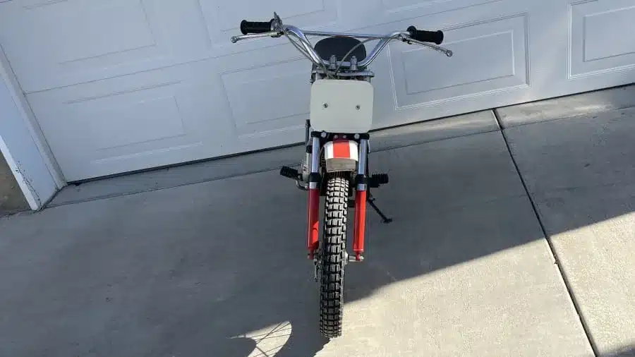 Frontansicht einer orangefarbenen 1971 Yamaha JT1 Mini Enduro mit Startnummernschild und grobstolligem Reifen, aufgenommen vor einer Garage.