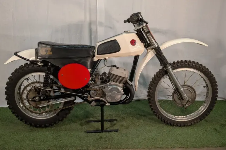 Seitenansicht einer 1973 CZ 125 Motocross mit weißem Tank, rotem Seitendeckel und Offroad-Bereifung.