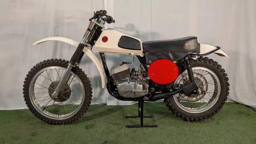 Seitenansicht einer originalen 1973 CZ 125 Motocross mit weißem Tank, rotem Rundschild und grobstolliger Offroad-Bereifung