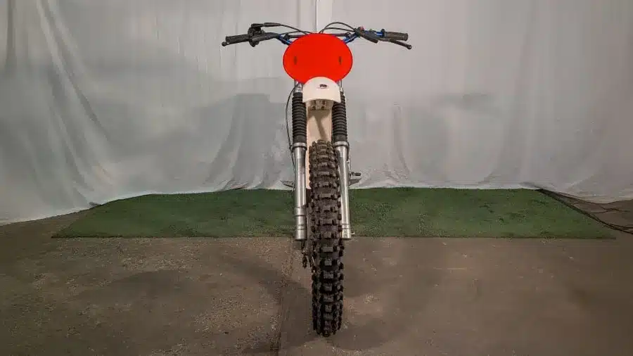 Frontansicht einer 1973 CZ 125 Motocross mit rotem Startnummernschild und grobstolliger Bereifung auf Betonboden vor neutralem Hintergrund.