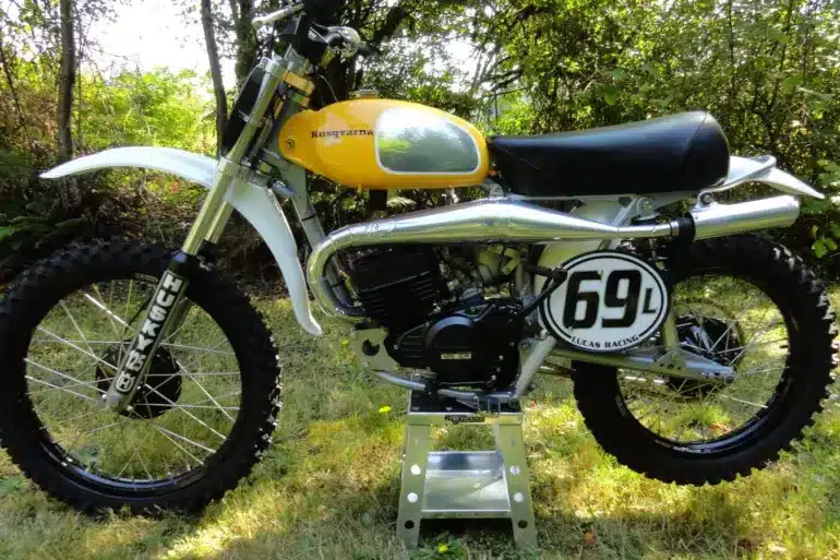 Seitenansicht einer Husqvarna 125CR Lightweight 1974 mit gelbem Tank, im Gelände auf einem Ständer platziert.