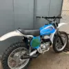 Restaurierte blaue Bultaco Pursang MK10 250cc von 1977, Seitenansicht vor grauer Wand.