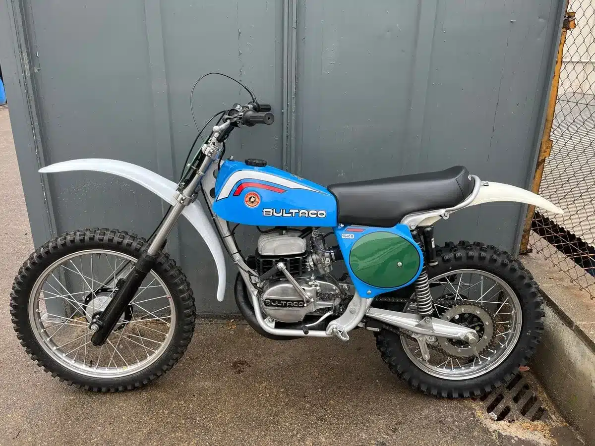 Seitenansicht einer restaurierten blauen Bultaco Pursang MK10 250cc von 1977 vor einer grauen Wand, klassische Motocross-Optik