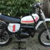 Seitliche Gesamtansicht einer 1977 Yamaha YZ80D Motocross-Maschine mit Startnummer 1, aufgestellt im Garten.