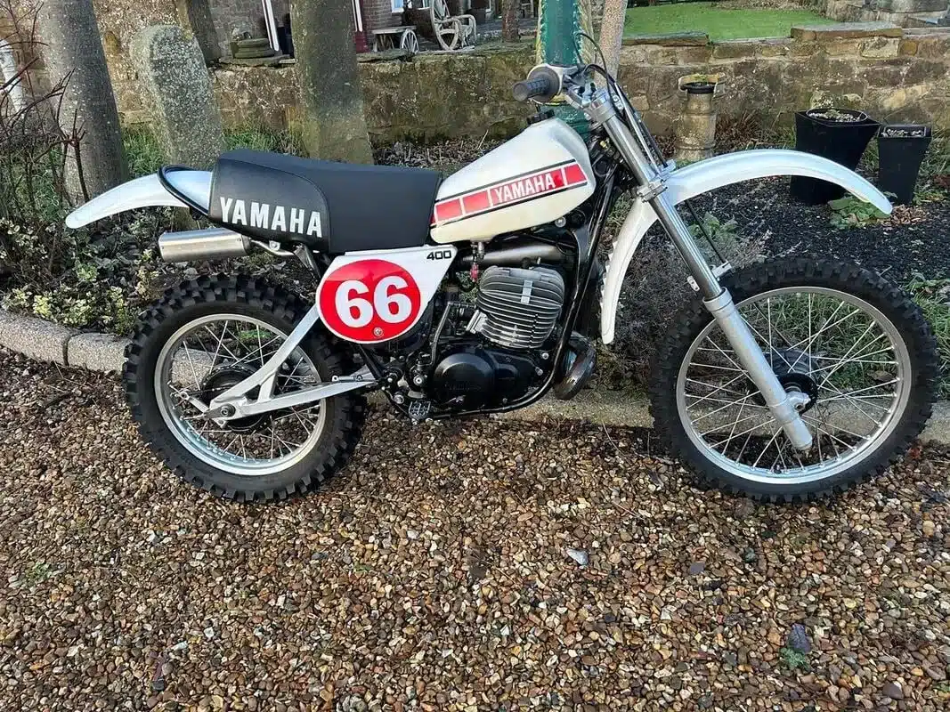 Seitenansicht einer restaurierten Yamaha YZ400E von 1978 mit Startnummer 66 und DG-Auspuff, geparkt auf Kies vor einer Steinmauer.