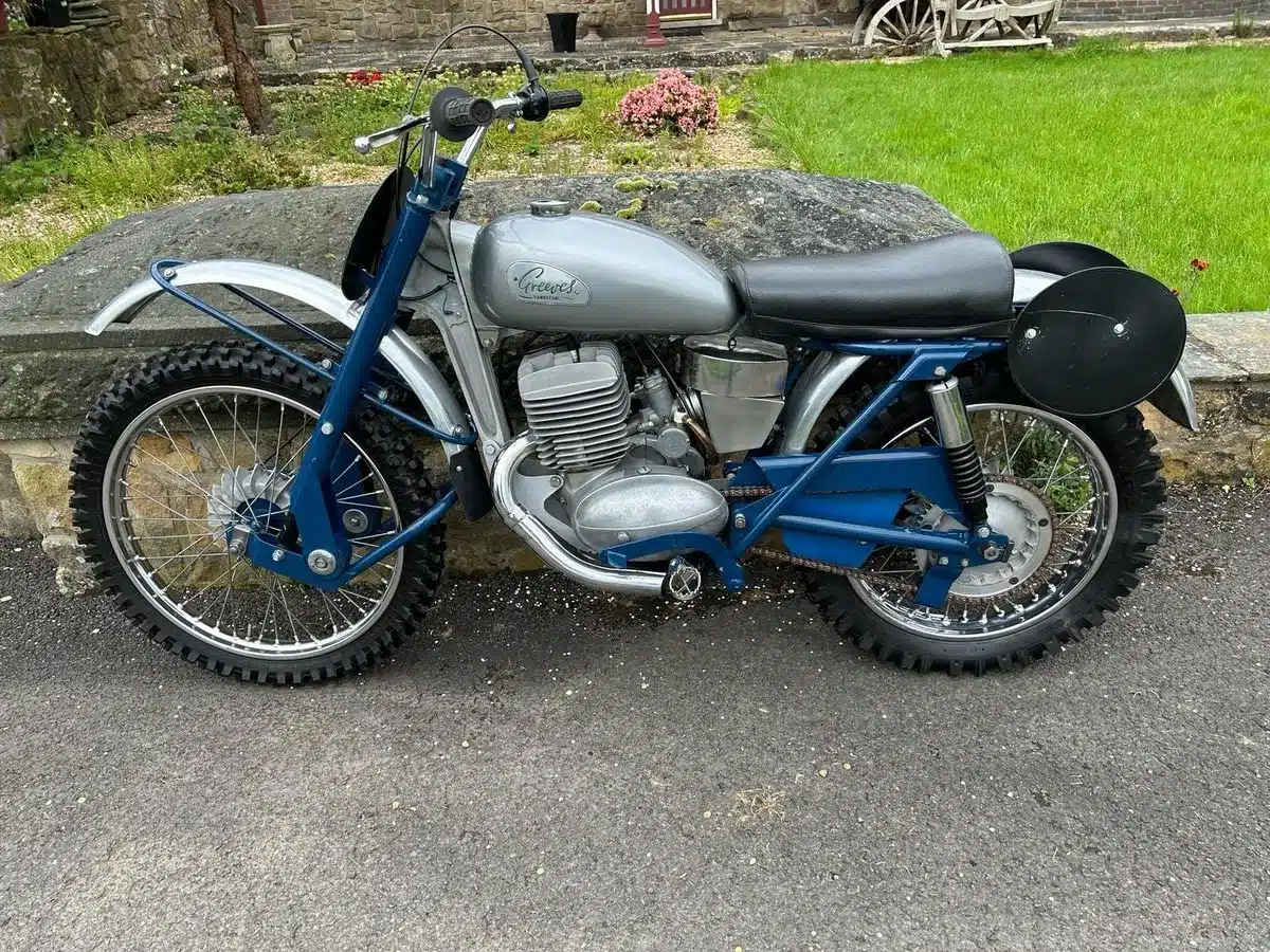 Eine restaurierte blaue Greeves Hawkstone MCS 250cc von 1960, abgestellt auf einer asphaltierten Fläche vor einer Steinmauer und Grünfläche.