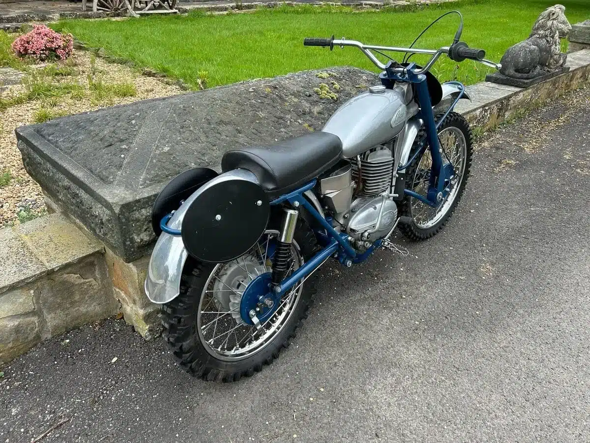 Seitliche Ansicht einer restaurierten blauen Greeves Hawkstone MCS 250cc von 1960 auf einem Asphaltweg, im Hintergrund Rasenfläche und Steinmauer