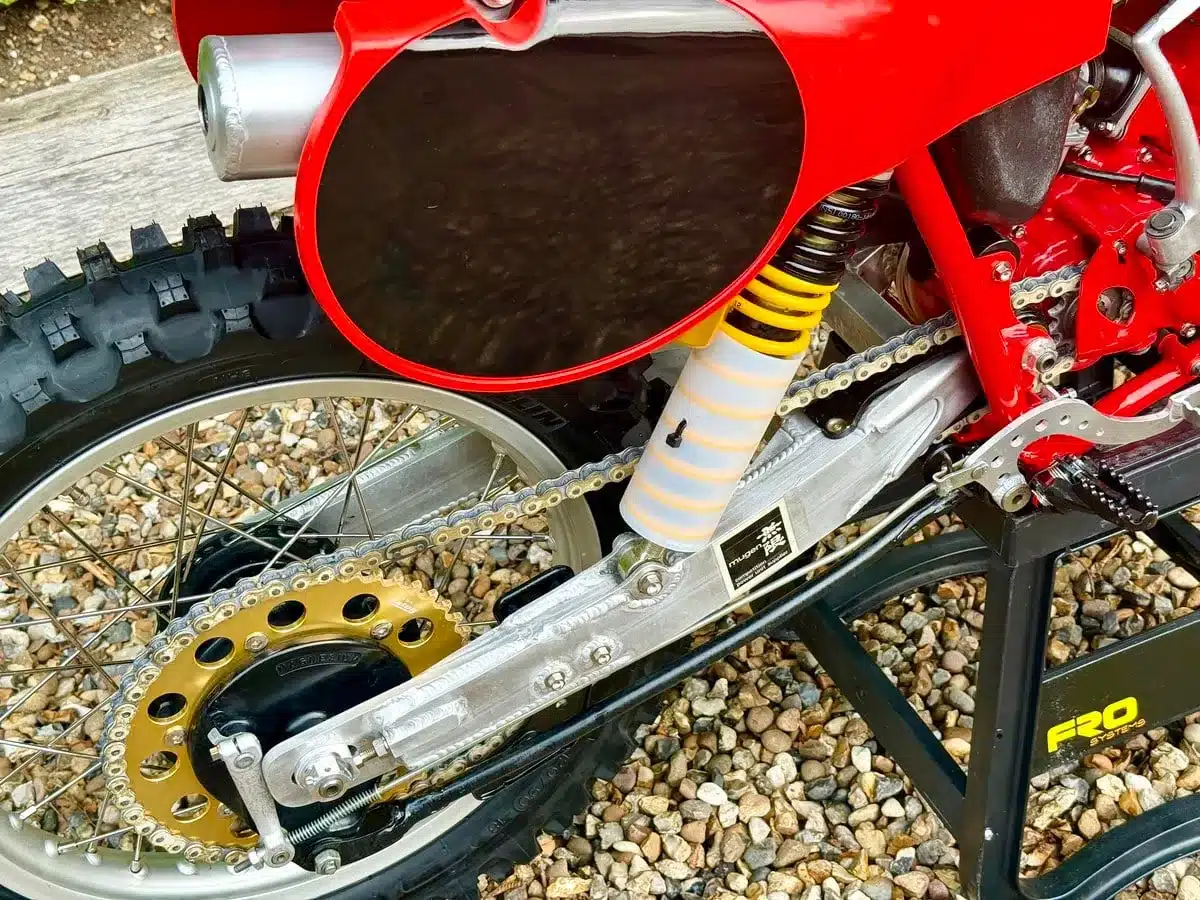 Detailaufnahme der Hinterradpartie einer Honda CR 125 mit Mugen-Banzai-Kit, sichtbare Alu-Schwinge, Öhlins-Federbein und goldene Antriebskette.