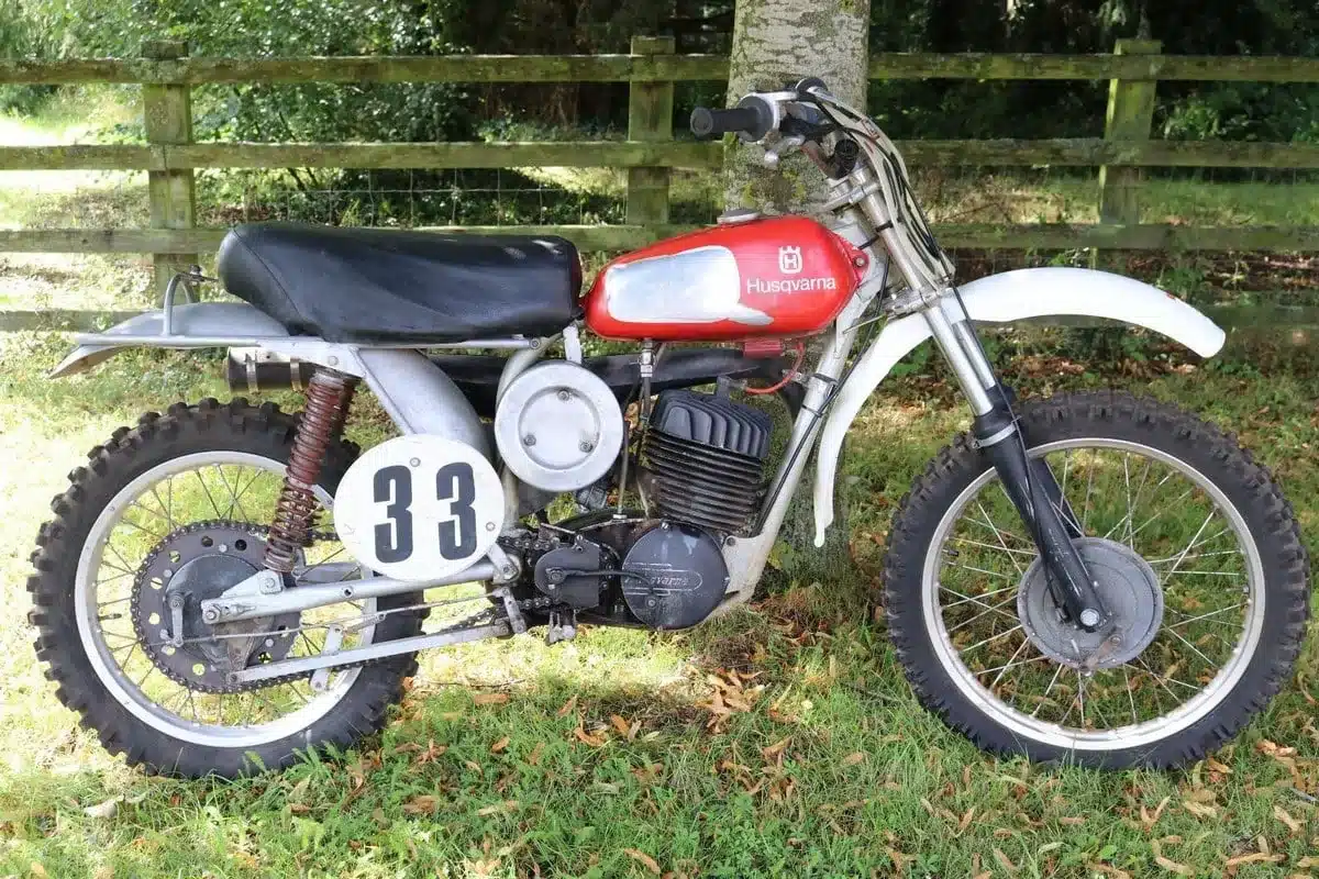 Seitenansicht einer Husqvarna 250 Cross von 1967 mit rotem Tank und Startnummer 33, platziert im Grünen vor einem Holzzaun.