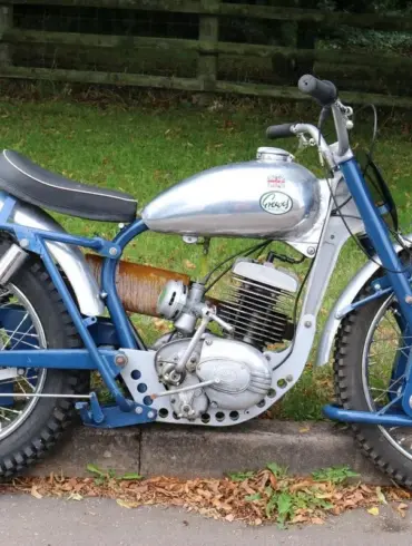 Trial-Motorrad mit gelbem Federbein, silbernem Tank und grobstolligen Reifen, geparkt am Straßenrand