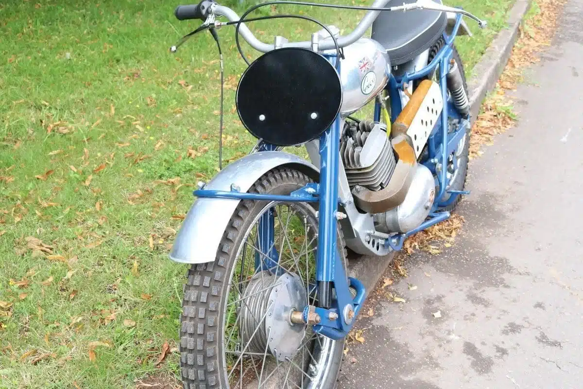 Altes britisches Motorrad mit blauem Rahmen und schwarzem Startnummernschild am Straßenrand, im Vordergrund das Vorderrad und Motordetails