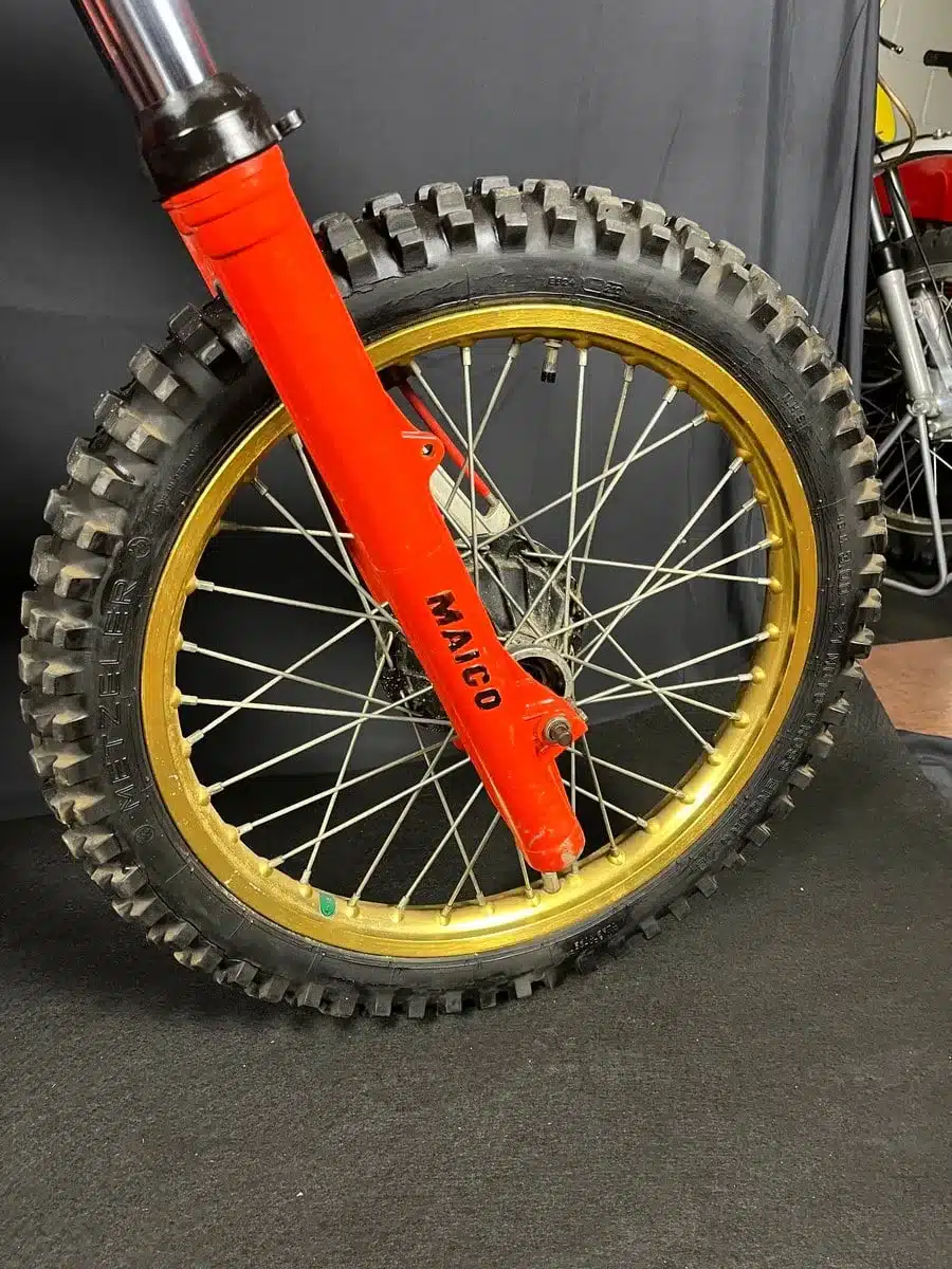 Nahaufnahme vom Vorderrad und Gabel einer Maico MC 250 von 1980 mit goldener Felge und grobstolligem Offroad-Reifen.