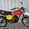 Montesa Enduro 250 von 1975, klassisches Enduro-Motorrad mit rotem Tank und gelben Startnummerntafeln seitlich, vor einer Metallwand abgestellt