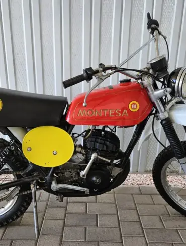 Montesa Enduro 250 von 1975, klassisches Enduro-Motorrad mit rotem Tank und gelben Startnummerntafeln seitlich, vor einer Metallwand abgestellt