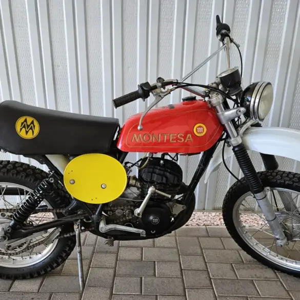 Montesa Enduro 250 von 1975, klassisches Enduro-Motorrad mit rotem Tank und gelben Startnummerntafeln seitlich, vor einer Metallwand abgestellt