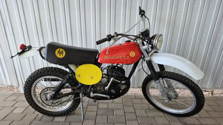 Montesa Enduro 250 von 1975, klassisches Enduro-Motorrad mit rotem Tank und gelben Startnummerntafeln seitlich, vor einer Metallwand abgestellt