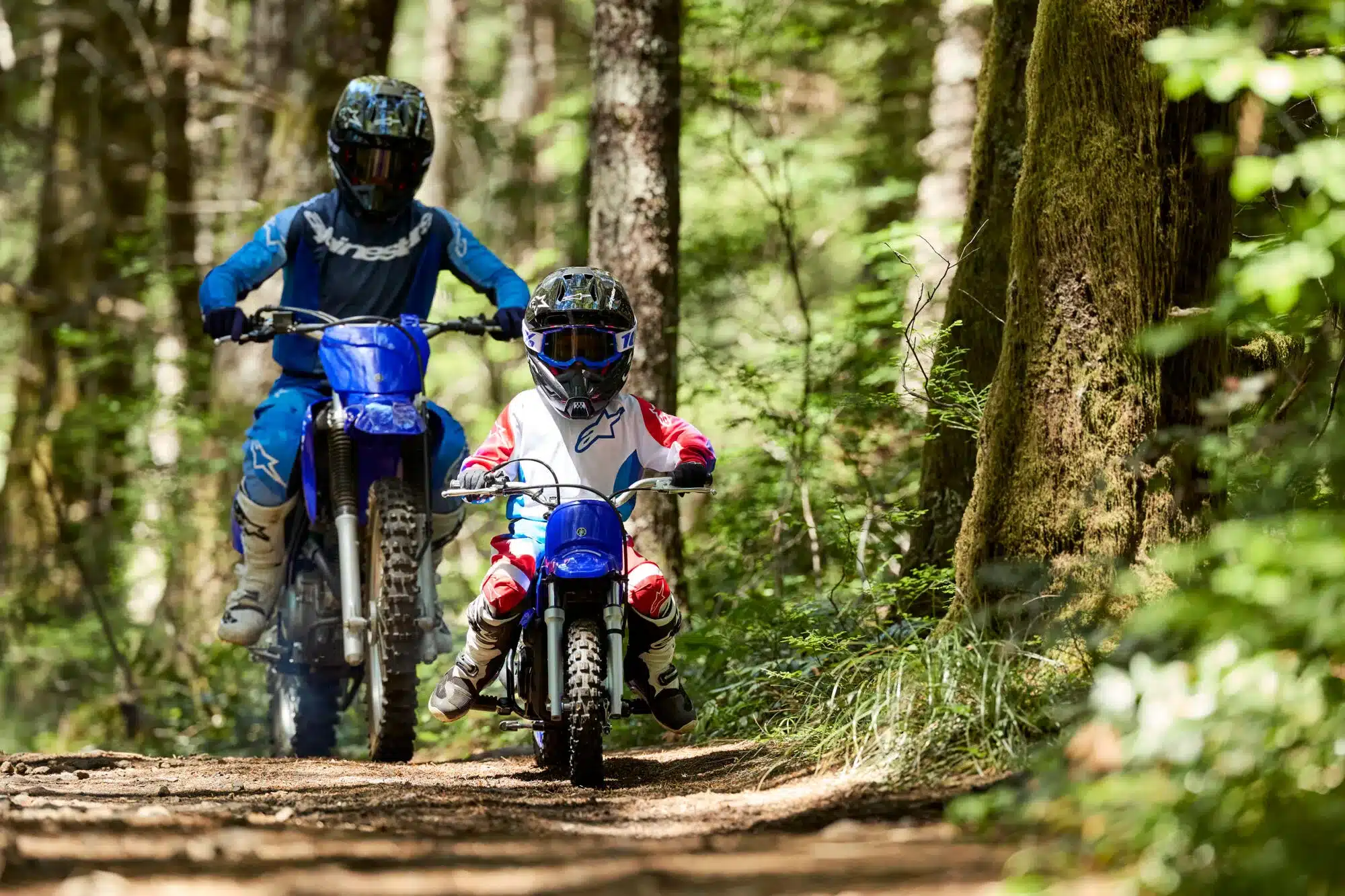 Ein Kind fährt im Wald auf einer blauen Yamaha PW50 vor einem Erwachsenen auf einem größeren Motorrad, beide in Motocross-Bekleidung.
