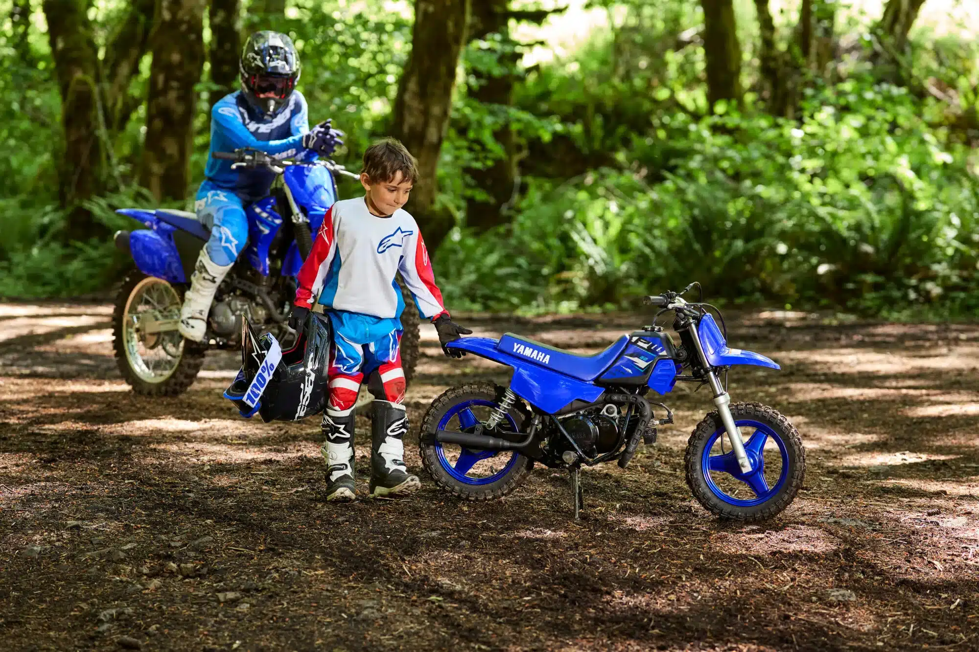 Kind in Motocross-Bekleidung neben einer Yamaha PW50 im Gelände, im Hintergrund Erwachsener auf größerem Motorrad