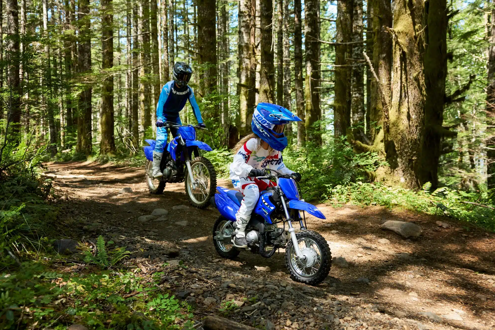 Kind auf einer blauen Yamaha TT-R50 fährt auf einem Waldweg vor einem Erwachsenen mit größerem Motorrad.