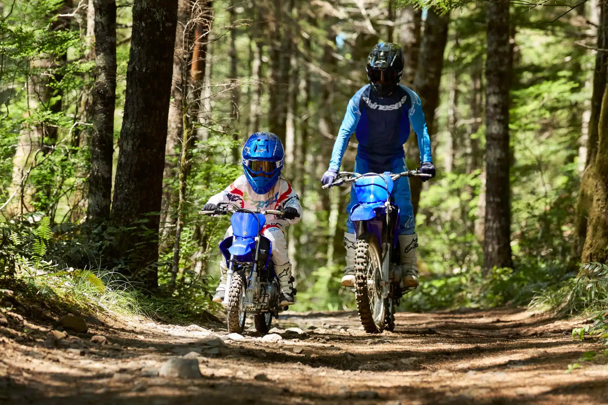 Kind fährt Yamaha TT-R50 und wird von Erwachsenem auf Yamaha-Motorrad auf einem Waldweg begleitet