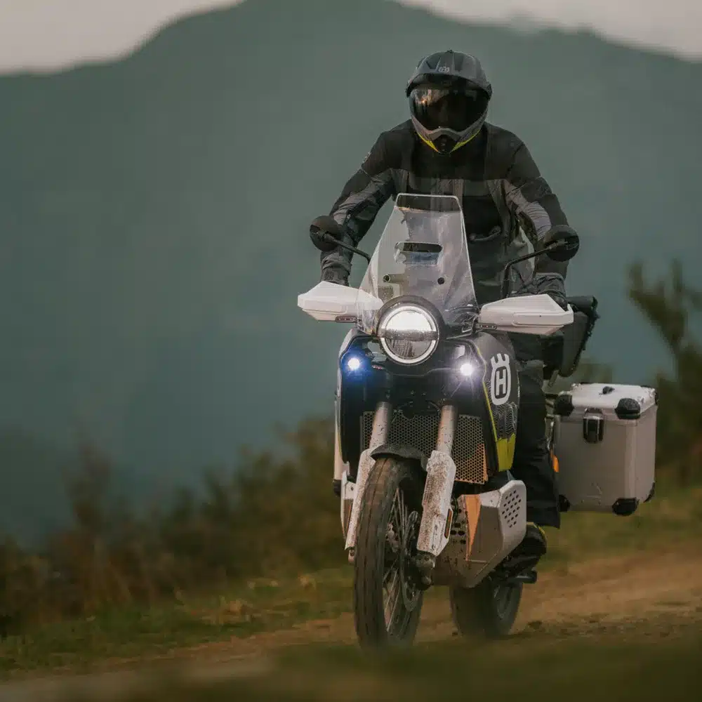 Fahrer auf einer Husqvarna Norden 901 Expedition 2026 fährt auf einer Schotterstraße im Gelände, umgeben von Bergen.
