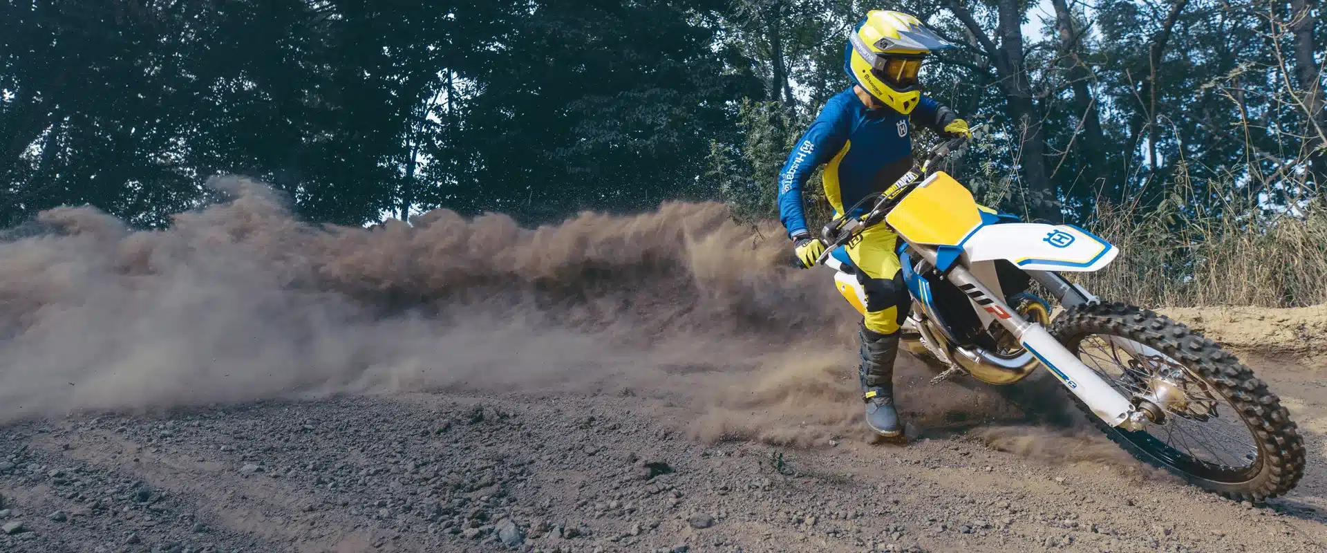 Fahrer auf einer Husqvarna TC 125 Heritage beim Driften auf einer Motocross-Strecke, umgeben von aufgewirbeltem Staub.