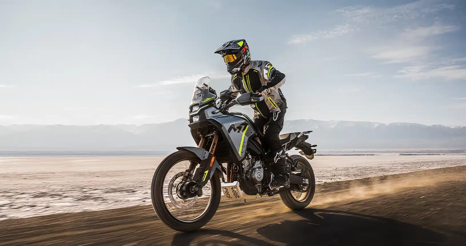 Fahrer auf einer CFMoto 450MT fährt dynamisch auf einer trockenen Schotterpiste vor Bergkulisse.