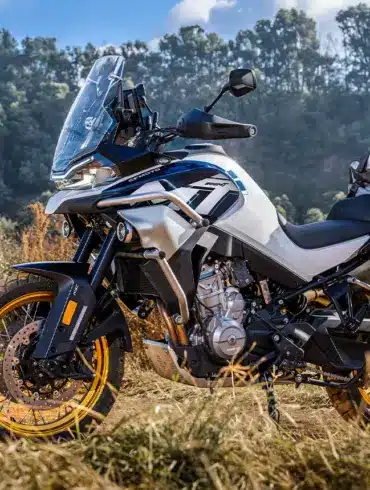 CFMOTO 800MT Explore Motorrad mit Gepäcktaschen auf Schotterweg und im Hintergrund bewaldete Hänge.