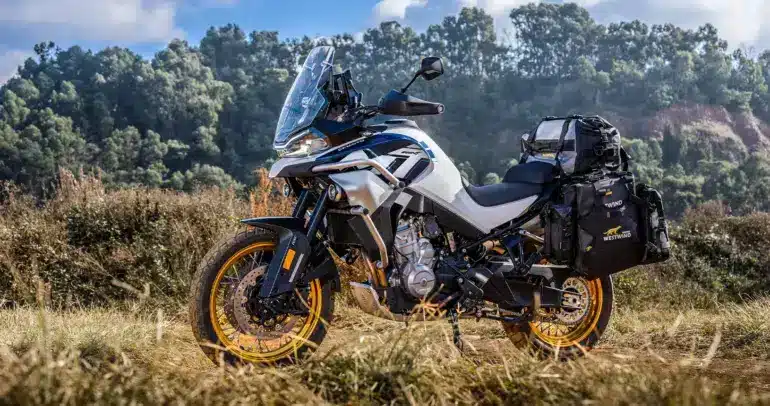 CFMOTO 800MT Explore Motorrad mit Gepäcktaschen auf Schotterweg und im Hintergrund bewaldete Hänge.