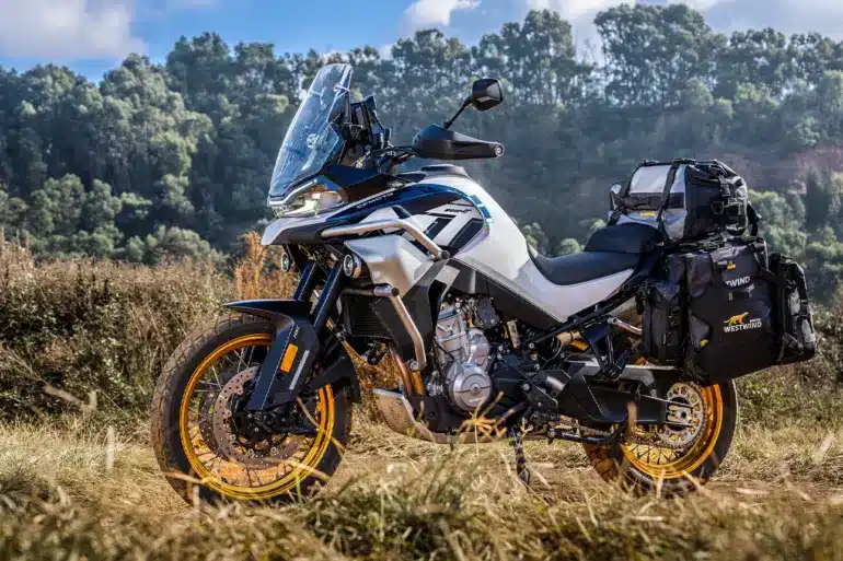 CFMOTO 800MT Explore Motorrad mit Gepäcktaschen auf Schotterweg und im Hintergrund bewaldete Hänge.