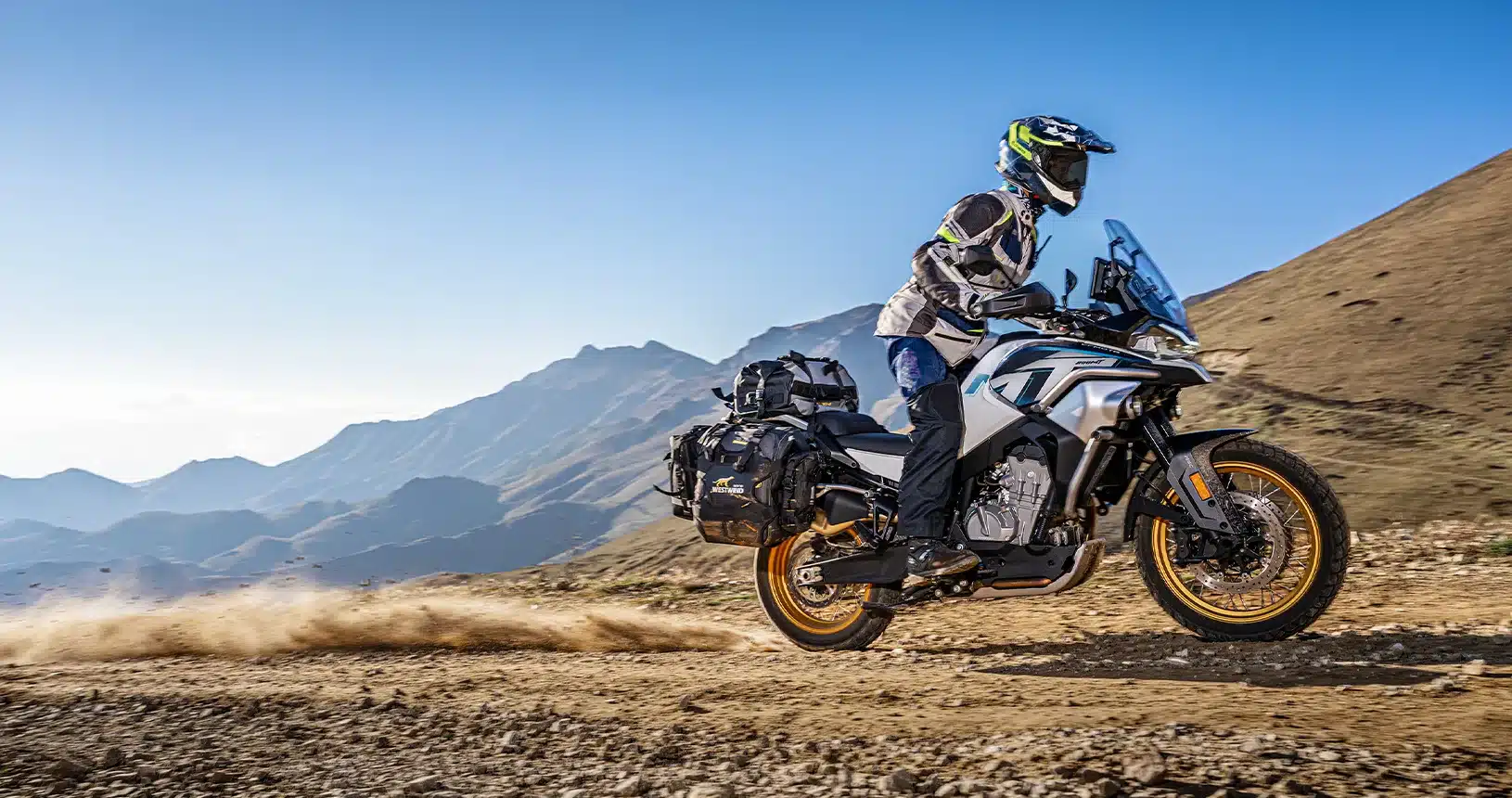 Abenteuer-Touring-Motorrad CFMOTO 800MT Explore mit Fahrer und Gepäck auf Schotterstraße vor Bergkulisse.