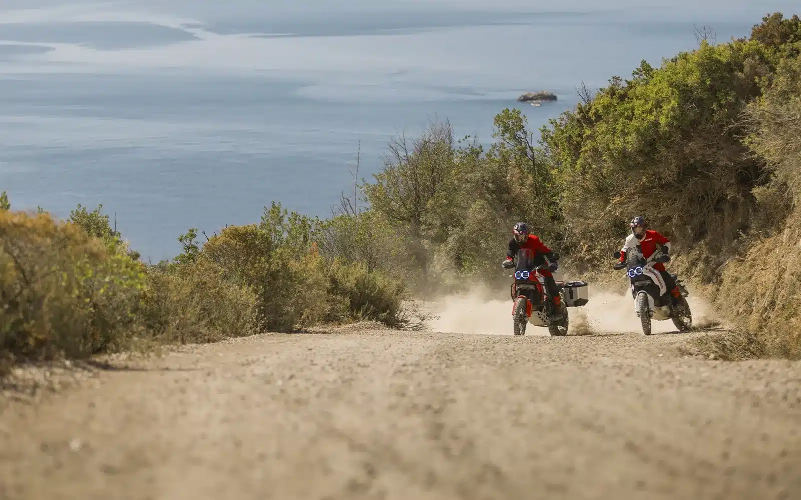Zwei Ducati DesertX Motorräder unterwegs auf einer Schotterpiste mit Meer im Hintergrund