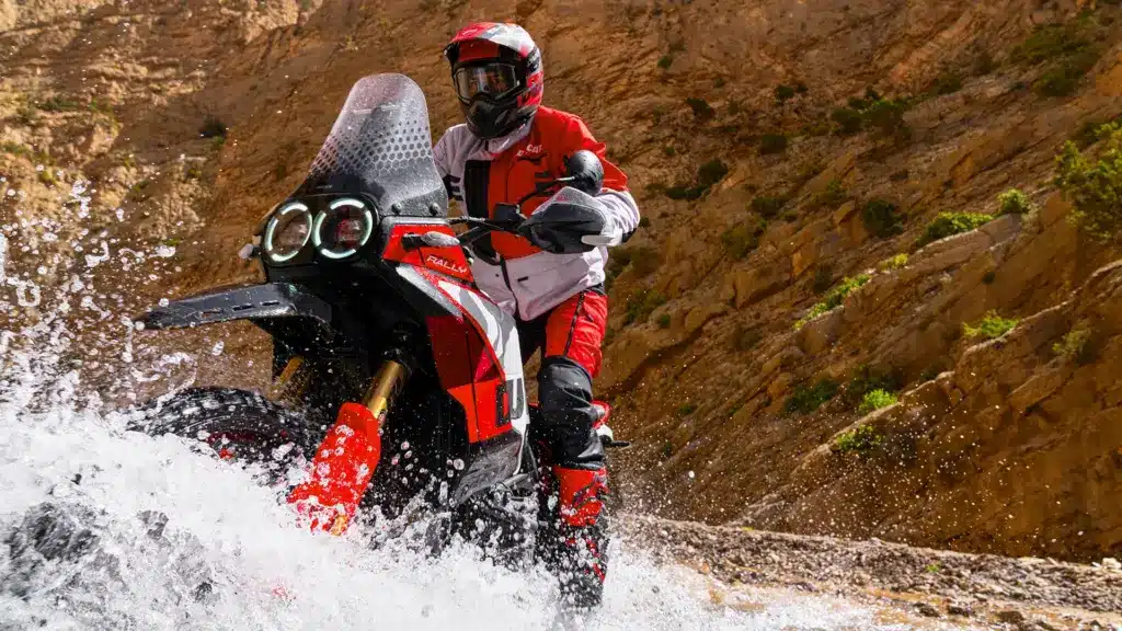 Ducati DesertX Rally fährt durch Wasser in einer Wüstenumgebung mit Felsen im Hintergrund.