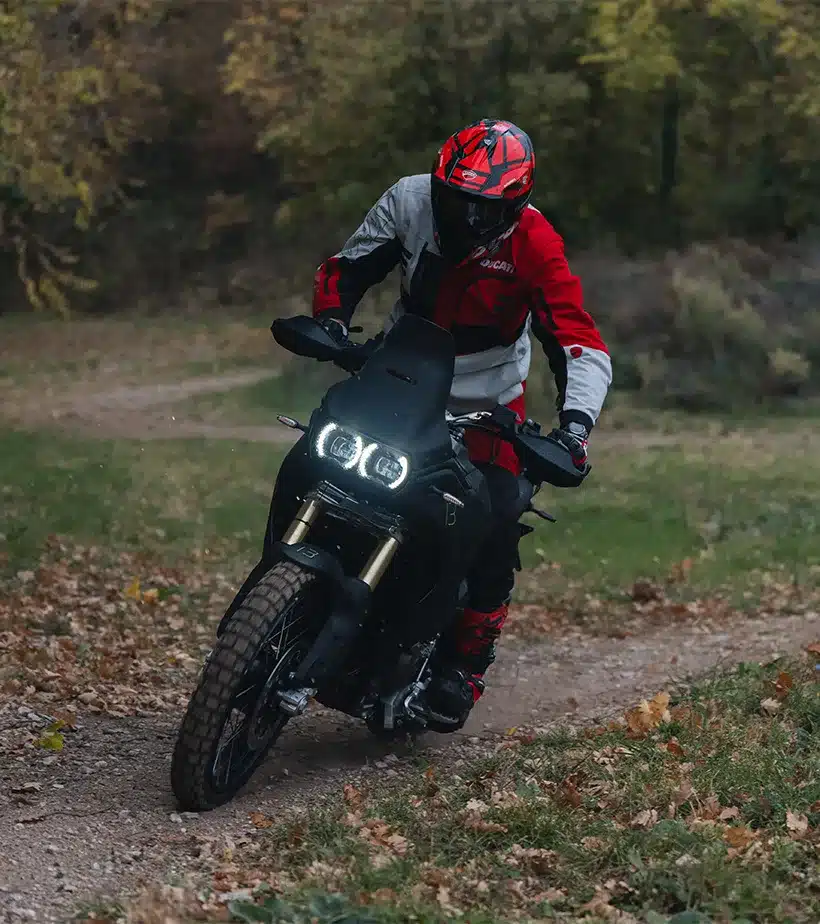 Ducati DesertX V2 fährt auf einem Schotterweg durch herbstliche Landschaft.