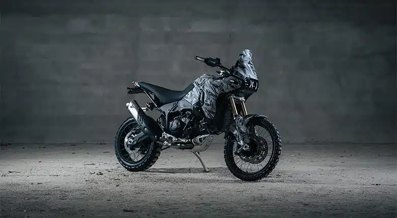 Getarnte Ducati DesertX V2, stehend auf Schotterboden vor grauer Wand