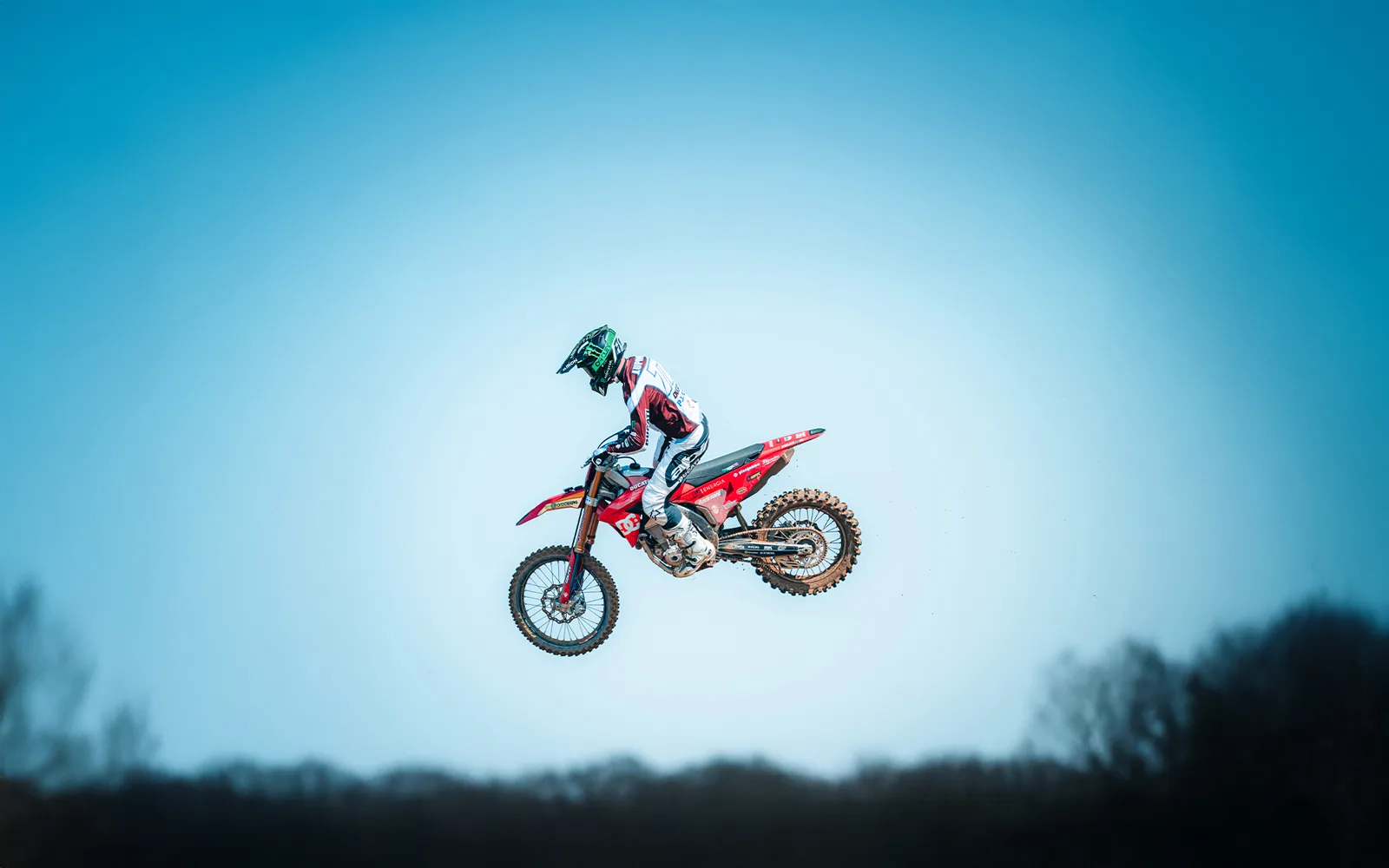 Ein Motocross-Fahrer mit Helm und Schutzkleidung springt mit einem roten Offroad-Motorrad vor blauem Himmel.