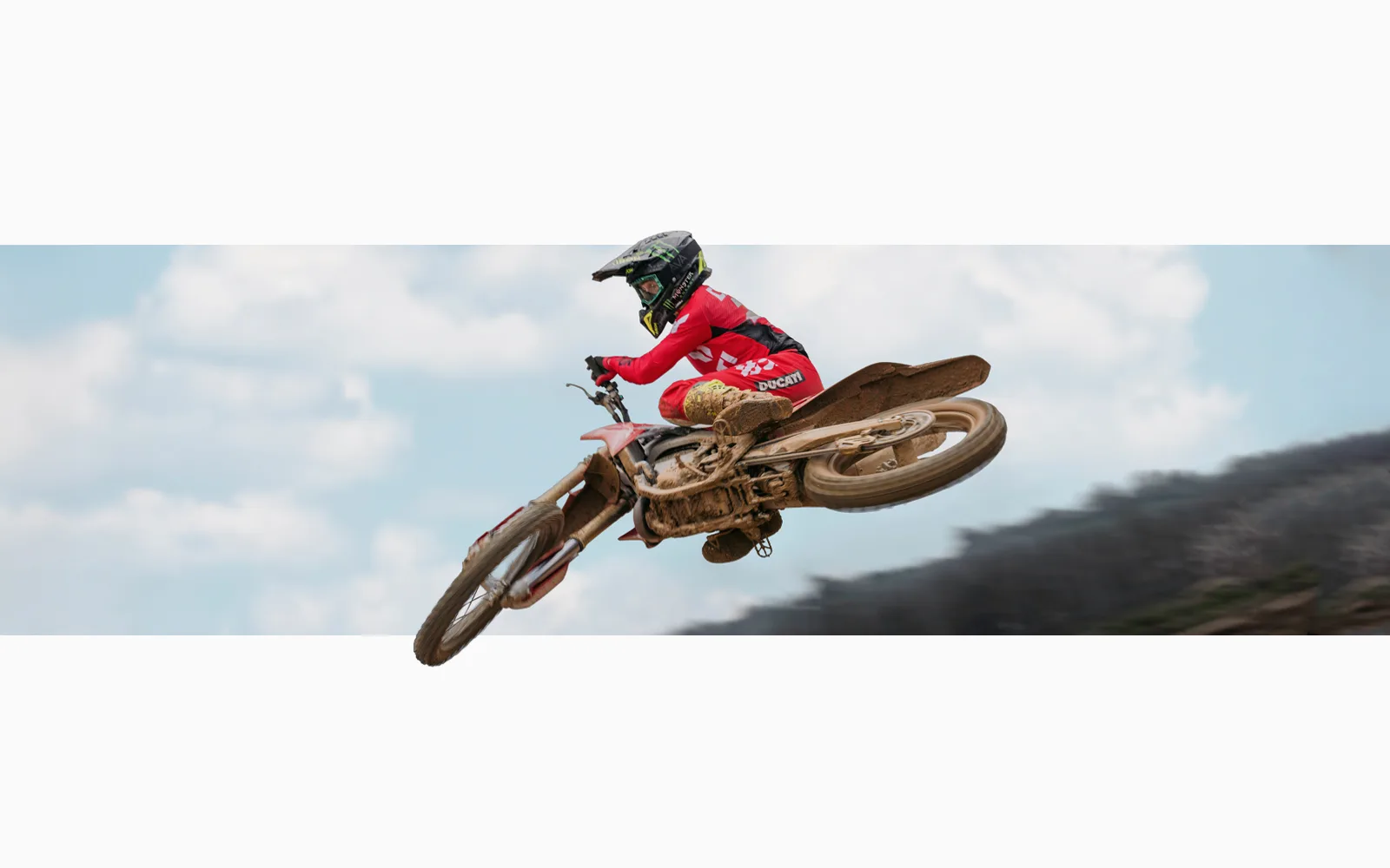 Motocross-Fahrer springt mit der Ducati Desmo450 MX über eine Strecke, Motorrad und Fahrer sind mit Schlamm bedeckt.