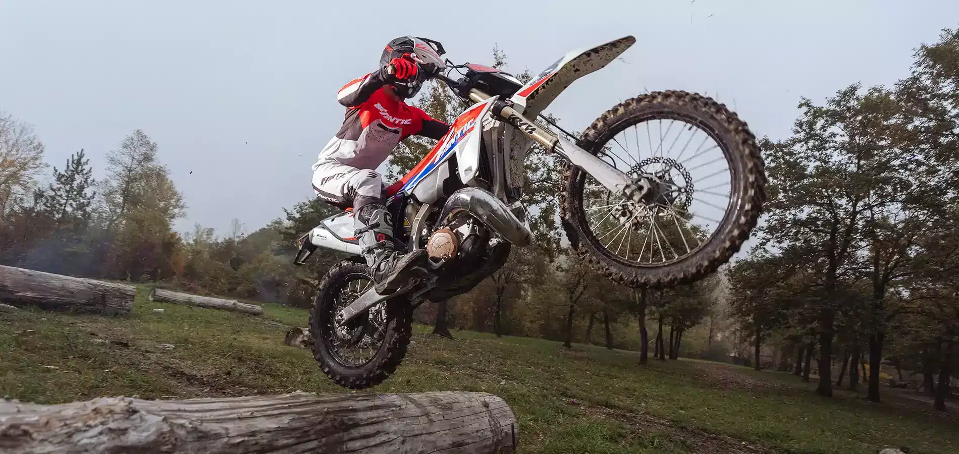 Fahrer springt mit einer Fantic XE 300 Enduro über einen Baumstamm im Wald.